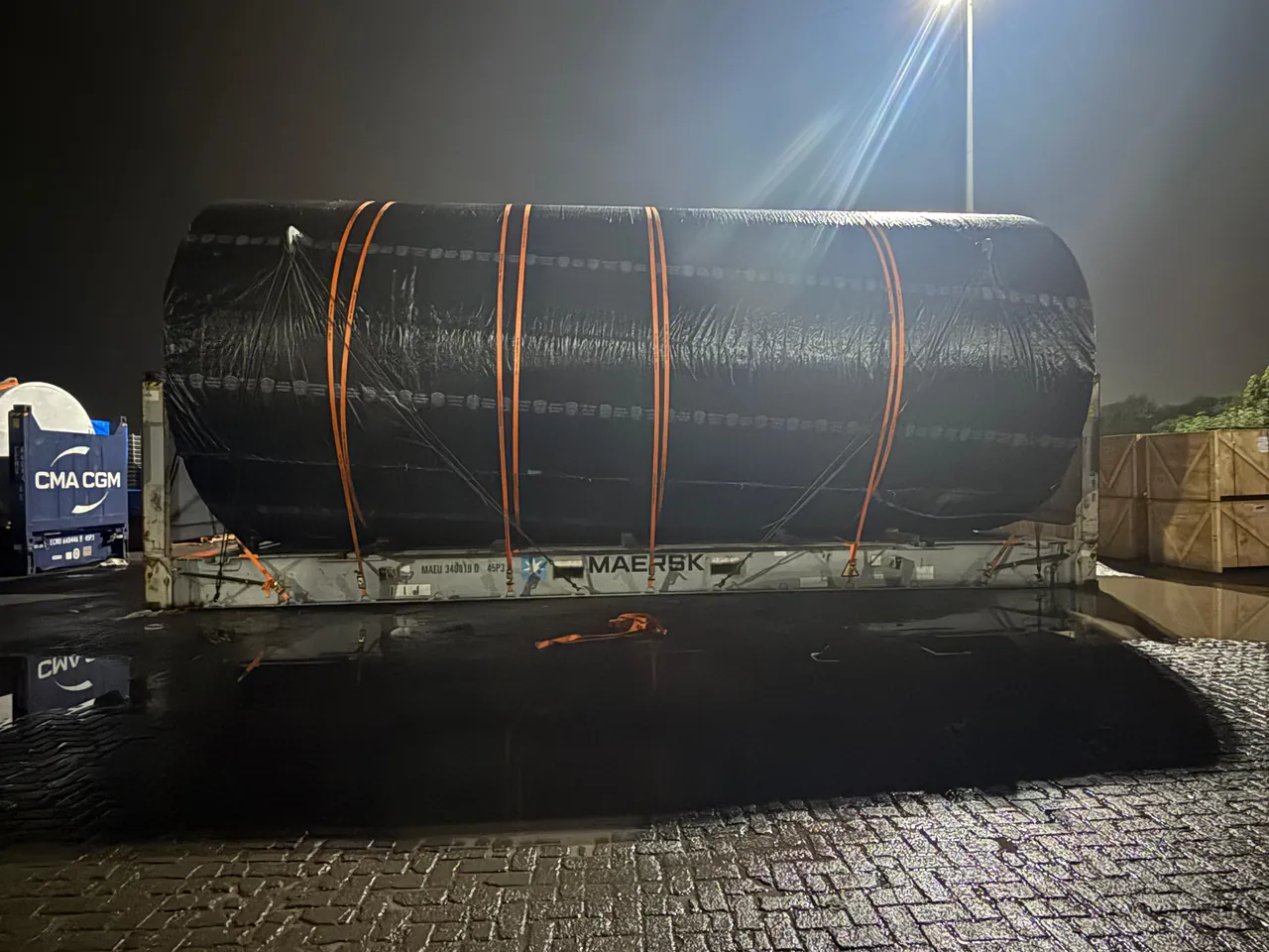 Large wrapped tank secured with lashing straps on a flat rack container at night